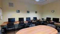 This single-floor computer lab is thoughtfully arranged to support both individual and collaborative work. The room features a long perimeter workstation desk lining the walls, accommodating multiple desktop computers complete with monitors, keyboards, and mice. A printer is also stationed along the desk, adding practical functionality for users. Each workstation is paired with a mobile, ergonomic chair, allowing for flexible seating arrangements. At the center of the room, a large rounded table provides additional space for group work or discussions, further enhancing the room's versatility. The walls are painted in a soft, neutral yellow-green tone that contributes to a calm and focused atmosphere. Framed artwork is displayed along the walls, adding a welcoming and personalized touch to the space. Overhead fluorescent lighting ensures consistent, even illumination throughout the room. The layout is clean and organized, with ample floor space allowing easy movement between workstations. Ventilation is provided via ceiling vents, supporting a comfortable working environment. Overall, this space is well-suited for use as a training room, classroom, or shared computer resource center.