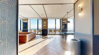 Located at 10-14 Lecount Place in New Rochelle, NY, this thoughtfully designed amenity lounge offers a sophisticated and versatile social environment across a single open floor. The space is anchored by striking herringbone-patterned hardwood floors that run throughout, lending warmth and visual interest to the entire area.

Floor-to-ceiling glass curtain walls along the perimeter flood the interior with abundant natural light and frame sweeping panoramic views of the sky and surrounding landscape, creating a bright, airy atmosphere. A bar-height counter with elegant cream-and-gold stools lines the window wall, offering an ideal setting for casual gatherings with a view.

The lounge features a bold, geometric blue-patterned accent wall that adds a vibrant, contemporary character to the space. A full-size pool table positioned beneath two industrial-style dome pendant lights serves as a central entertainment focal point. Adjacent to it, a richly upholstered tan leather sectional sofa faces a wall-mounted flat-screen TV, creating a comfortable media and lounging area.

A semi-private nook with frosted glass partitions provides quieter seating options, furnished with modern white chairs and a sleek dark work surface — ideal for focused conversation or casual work. Warm sconce lighting and recessed ceiling fixtures complete the polished, modern aesthetic throughout.