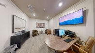 Located at 4947 Stewart Mill Rd in Douglasville, Georgia, this single-room professional space is thoughtfully designed to serve both as a conference room and a comfortable lounge area. The room features crisp white walls with subtle paneling details and neutral-toned carpet flooring, creating a clean and polished atmosphere throughout.

The primary workspace centers around a round wood-toned conference table surrounded by tan leather chairs with gold-accented bases, equipped with a desktop computer, printer, and keyboard setup for productive meetings. A large wall-mounted flat-screen TV is positioned above the workstation wall, ideal for presentations or video conferencing.

On the opposite side of the room, a relaxed lounge area provides a welcoming contrast, featuring two olive-green upholstered armchairs arranged around a small side table. A dark wood credenza along the adjacent wall displays decorative accents and is topped with framed artwork, adding warmth and character to the space. A second piece of wall art — a vibrant floral painting — anchors the lounge corner with a touch of color.

Recessed ceiling lighting provides even illumination throughout, while a glass-paneled green door offers a stylish entry point. The overall ambiance is professional yet inviting, making it well-suited for small meetings, client consultations, or collaborative work sessions.