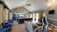 This well-appointed single-floor community clubhouse offers a thoughtfully designed gathering space that balances comfort and functionality. The open-concept layout is anchored by soaring vaulted ceilings, which are accentuated by a ceiling fan and recessed lighting, creating a bright and airy atmosphere throughout.

The lounge area is a central highlight, featuring a striking floor-to-ceiling tile fireplace with a classic hearth surround, a wall-mounted flat-screen television, and a vibrant geometric area rug. Comfortable seating arrangements, including plush sofas and accent chairs in neutral tones, are accented with bold teal, blue, and yellow decorative pillows that complement the space's color palette.

Adjacent to the lounge is a wet bar and peninsula with a white quartz countertop, mosaic tile base, and bar seating with blue-cushioned chairs — ideal for casual gatherings. A pass-through window connects this area to a kitchenette behind, and large glass-panel walls provide a direct view into the on-site fitness center equipped with exercise machines.

Multiple French doors and large windows dressed in coordinating yellow-and-navy curtains flood the space with natural light and offer views of the surrounding landscaped exterior. A café-style seating area near the windows provides additional space for relaxing or socializing. Decorative artwork and sculptural accents complete the polished, contemporary interior design.