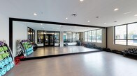 This single-level fitness studio is designed to accommodate a wide range of exercise activities within a clean, modern environment. The most prominent feature is the expansive wall of floor-to-ceiling mirrors framed in sleek black, which lines the main exercise floor and visually doubles the sense of space while allowing users to monitor their form during workouts. Recessed ceiling lighting provides bright, even illumination throughout the room, complementing the natural light that filters in through large windows along the perimeter wall, offering views of lush greenery outside.

The open studio floor is finished with smooth, light-toned flooring ideal for group fitness classes, yoga, stretching, or cardio activities. Along the side walls, a dumbbell rack stocked with a range of free weights provides convenient access for strength training. Colorful resistance bands and additional equipment are neatly stored nearby. Black-framed glass doors connect the studio to an adjacent fitness area visible in the mirror's reflection, which appears to house additional cardio and strength training machines, making the overall facility well-equipped for a comprehensive workout experience.