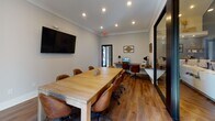 This single-floor workspace is thoughtfully designed to accommodate a variety of professional needs, combining a conference area with dedicated individual workstations in a cohesive, modern environment. The space features warm hardwood-style flooring throughout, crisp white walls with crown molding, and recessed lighting that creates a bright, welcoming atmosphere.

The central conference area is anchored by a substantial light wood rectangular table surrounded by leather rolling chairs, providing ample seating for group meetings or collaborative sessions. A wall-mounted flat-screen television serves as the primary display for presentations. Natural light enters through a large window dressed with patterned curtains, while decorative black metal wall panels add visual interest. A floor-to-ceiling glass partition with a black frame elegantly separates the conference space from the adjacent workstation area, maintaining an open feel while defining distinct zones.

The dedicated workstation nook features an L-shaped floating desk with two computer setups, each accompanied by a leather task chair and individual desk lamp. Decorative framed wall art completes the area. A glass door provides access to an outdoor area, further enhancing the sense of connectivity and airflow throughout the space.