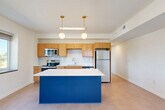 Located at 8 S Sixth St in Council Bluffs, Iowa, this one-bedroom residence offers a thoughtfully designed single-level layout. A clean, well-lit corridor with recessed lighting and crisp white walls leads to the main living areas, establishing a bright and modern aesthetic throughout.

The kitchen is a standout feature, showcasing warm honey-toned cabinetry, white subway tile backsplash, and a full suite of stainless steel appliances including a range, microwave, and refrigerator. A striking cobalt blue island with a white countertop serves as the kitchen's centerpiece, complemented by elegant brass pendant lighting overhead. A window above the kitchen area frames an outdoor view, filling the space with natural light.

The generously sized primary bedroom offers a quiet retreat, featuring plush carpeting, recessed lighting, and a window overlooking the surrounding streetscape. Multiple doorways provide access to closet space and additional rooms, offering practical storage and organization.

The dedicated laundry area is a convenient addition, housing a stacked washer and dryer unit within its own alcove, keeping household functions neatly contained. Throughout the residence, a consistent palette of white walls, light flooring, and clean lines creates a cohesive and modern living environment.