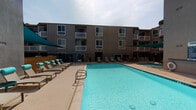 This inviting outdoor amenity area is designed for relaxation and social gathering, centered around a generously sized swimming pool with crystal-clear turquoise water. The pool deck is lined with cushioned lounge chairs and accented by vibrant teal shade sails that provide comfortable relief from the sun, creating a resort-like atmosphere.

Adjacent to the pool, a dedicated outdoor lounge and dining area offers a fully equipped outdoor kitchen complete with a stainless steel BBQ grill, granite countertops, and built-in storage cabinetry, all set against a warm wood-paneled backdrop. A large metal dining table with seating for several guests makes this space ideal for entertaining. Lush greenery panels line the perimeter fencing, adding a touch of privacy and natural texture to the setting.

The entire space is enclosed by a black iron security gate, ensuring a secure and private environment. The surrounding multi-story building features numerous private balconies that overlook the courtyard, enhancing the sense of a connected community. The open sky above allows for abundant natural light throughout the day, making this outdoor space a standout amenity for relaxation, recreation, and social gatherings.