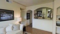 This single-floor residence showcases a thoughtfully designed interior that balances functionality with classic architectural detail. The living area is anchored by warm hardwood-style flooring that extends throughout, creating a cohesive and inviting atmosphere. Elegant arched doorways serve as a defining architectural feature, lending a sense of sophistication and flow between rooms. Crown molding along the ceilings adds a refined finishing touch that elevates the overall aesthetic.

A standout feature of the space is the built-in arched pass-through connecting the living area to the kitchen, complete with a light-colored stone countertop and integrated display shelving. This design element promotes an open feel while maintaining a subtle separation between spaces. The shelving within the archway provides both decorative and practical storage opportunities.

The living room is furnished with comfortable seating and accented by a classic table lamp that contributes to the warm, welcoming ambiance. Neutral wall tones throughout create a versatile backdrop, allowing the architectural details to take center stage. Arched passageways lead to additional rooms, reinforcing the home's consistent design language. Overall, the space presents a harmonious blend of classic architectural elements and practical living comfort.