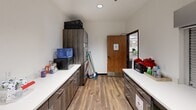 Located in Phoenix, Arizona, this single-floor community room is a generously sized, multipurpose gathering space designed to serve a diverse range of activities and needs. The open main area features warm wood-look flooring, a drop-panel ceiling with ample overhead lighting, and a layout that accommodates multiple functional zones simultaneously. Dining and activity tables with chairs are arranged throughout, providing space for group meals, events, and children's activities, while a comfortable seating area with sofas offers a more relaxed environment for casual gatherings. A dedicated computer station area lines one wall, equipped with adjustable desks and task chairs, framed by botanical artwork that adds a fresh, welcoming aesthetic. Decorative touches including indoor trees, wall clocks, framed artwork, and a children's reading nook contribute to the room's warm and community-oriented atmosphere. A "Gather" sign and colorful displays reinforce the space's inviting, inclusive character. The kitchen is outfitted with dark wood cabinetry, white countertops, a full-size refrigerator, and a microwave, supporting food preparation and storage for events. Additional storage and utility areas are accessible off the main space. Throughout, the design balances practicality with a comfortable, community-focused ambiance well suited for a variety of programs and gatherings.