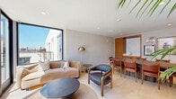 Located at 1430 Lincoln Blvd in the heart of Santa Monica, California, this thoughtfully designed single-floor space blends modern aesthetics with warm, functional details. The living room serves as the centerpiece, bathed in abundant natural light streaming through expansive floor-to-ceiling windows framed in sleek black. A sliding glass door opens directly to an outdoor terrace, creating a seamless indoor-outdoor connection and offering bright California views.

The space features light-toned hardwood flooring throughout, lending warmth and continuity. A comfortable seating arrangement with a curved beige sofa, a sculpted dark round coffee table, and a leather accent chair creates an inviting lounge area, complemented by a brass floor lamp and an area rug with a geometric pattern.

A large oval conference table surrounded by leather dining chairs defines a dedicated meeting or dining zone, accented by a curated gallery wall of framed artwork and a wall-mounted display screen. Warm wood cabinetry with a marble-topped wet bar adds a refined, functional touch to this area.

A tall potted palm brings a natural, tropical element to the space, reinforcing the bright and airy Southern California atmosphere. Recessed lighting, clean white walls, and contemporary furnishings complete this polished, versatile space.