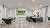 This single-floor community clubhouse offers a well-appointed and versatile layout designed to serve both social and administrative functions. The entrance greets visitors with light wood-look tile flooring, sheer linen curtains, and an abundance of natural light streaming through large glass double doors and surrounding windows. A small seating area and dining table near the entry provide an inviting welcome space.

The open lounge and living area is a standout feature, boasting soaring vaulted ceilings accented by exposed wooden beams and a striking floor-to-ceiling natural stone fireplace. This area is furnished with plush green velvet sofas, a shuffleboard table, and warm, layered décor that creates a relaxed, residential ambiance. French doors lead to an outdoor view of what appears to be a pool area, further enhancing the bright and airy atmosphere.

The leasing office section features a large dark-toned desk with upholstered green chairs, a circular mirror, and elegant pendant lighting. A dedicated service counter along the back wall is finished with bold green hexagonal tile, white cabinetry, and pendant lights — functioning as a refreshment station.

The fully equipped kitchen includes white shaker-style cabinetry with matte black hardware, a textured tile backsplash, light quartz countertops, and stainless steel appliances, including a refrigerator, range, and dishwasher. Elevated ceilings carry through this space as well, maintaining the open, expansive feel throughout.