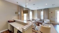 This single-floor community room offers a welcoming and sophisticated gathering space designed with both comfort and style in mind. At the forefront, a substantial granite-topped bar area with crisp white cabinetry serves as a functional centerpiece, ideal for hosting and socializing. Overhead, a pair of brushed gold drum pendant lights add a refined, contemporary touch, complementing the warm neutral palette found throughout the space.

The open lounge area beyond features plush, light-toned seating arrangements centered around a wood-accented coffee table, creating an inviting environment for casual conversation or relaxed gatherings. Hardwood flooring anchors the space with warmth, while large windows dressed in floor-length linen drapes allow generous natural light to fill the room. A set of glass doors at the rear further enhances the sense of openness and connectivity to the outdoors.

Recessed lighting supplements the natural light, ensuring the space remains bright and comfortable throughout the day. The overall ambiance is elegant yet approachable, with a cohesive design language of soft neutrals, metallic accents, and quality finishes throughout.