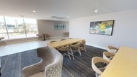This contemporary single-floor communal space is thoughtfully designed to accommodate both collaborative gatherings and relaxed, informal interaction. The open layout flows seamlessly across distinct seating zones, each catering to a different social dynamic. A central area features a long, light wood communal table surrounded by modern chairs, ideal for group meetings or shared work sessions. Adjacent to this, a lounge zone offers curved, upholstered seating and stylish wingback chairs arranged for comfortable conversation. The light-toned hardwood flooring transitions to a patterned, dark-hued carpet with gold accent lines, visually anchoring the various functional areas throughout the space. Recessed lighting provides consistent, even illumination across the interior, complemented generously by natural light streaming through expansive floor-to-ceiling windows along one side of the building. These large windows frame outdoor views and create a bright, airy atmosphere throughout. A dedicated casual seating nook near the windows features compact bistro-style tables and chairs, perfect for smaller gatherings. The walls are painted in a clean white and soft grey palette, accented by vibrant abstract artwork in teal, yellow, and blue tones, adding a contemporary artistic flair. The overall ambiance is clean, modern, and welcoming, well-suited for community use.