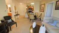 This single-floor residence showcases an open-concept layout that integrates the living, dining, and kitchen areas into one cohesive, flowing space. The living area features plush wall-to-wall carpeting in a warm neutral tone, providing a comfortable foundation for the stylishly furnished seating arrangement, which includes a sofa accented with decorative pillows and a gold-toned circular coffee table. Adjacent to the living area, a round dining table with dark, curved-back chairs creates an intimate dining setting, positioned naturally between the living space and kitchen. The kitchen is defined by warm wood cabinetry, black appliances including a microwave and range, and a peninsula with a stone-style countertop that serves as both a prep area and a casual divider between the kitchen and dining zone. Pendant lighting hangs above the peninsula, adding a decorative touch while enhancing task lighting. A decorative console table near the entryway, paired with wall art and floral accents, adds character to the foyer transition. The warm, ambient lighting throughout contributes to an inviting and well-appointed atmosphere.