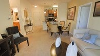 This single-floor residence showcases an open-concept layout that integrates the living, dining, and kitchen areas into one cohesive, flowing space. The living area features plush wall-to-wall carpeting in a warm neutral tone, providing a comfortable foundation for the stylishly furnished seating arrangement, which includes a sofa accented with decorative pillows and a gold-toned circular coffee table. Adjacent to the living area, a round dining table with dark, curved-back chairs creates an intimate dining setting, positioned naturally between the living space and kitchen. The kitchen is defined by warm wood cabinetry, black appliances including a microwave and range, and a peninsula with a stone-style countertop that serves as both a prep area and a casual divider between the kitchen and dining zone. Pendant lighting hangs above the peninsula, adding a decorative touch while enhancing task lighting. A decorative console table near the entryway, paired with wall art and floral accents, adds character to the foyer transition. The warm, ambient lighting throughout contributes to an inviting and well-appointed atmosphere.