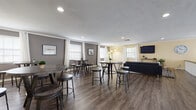 Located at 1343 Weathervane Ln in Akron, Ohio, this single-floor community facility offers a well-organized and thoughtfully designed layout suited for gathering, relaxing, and communal activities. The entrance greets visitors with a reception area featuring a white brick-accented counter, warm wood double doors, and durable luxury vinyl plank flooring that carries throughout much of the space. Adjacent to the entry, an open dining area provides multiple high-top round tables with bar stools, bathed in natural light from several windows dressed with white curtains and blinds. Neutral gray and yellow tones on the walls lend a calm, welcoming atmosphere. The lounge area flows naturally from the dining space and includes a large sectional sofa, a wall-mounted flat-screen television, and greenery that adds a refreshing touch. A standout feature of the space is a dedicated sitting room centered around a striking freestanding circular fireplace with a conical metal hood, set on a tiled raised hearth and surrounded by plush seating — creating a cozy and distinctive focal point. The kitchen is fully equipped with wood cabinetry, standard appliances including a refrigerator, stove, dishwasher, and microwave, and offers ample counter space for food preparation. Recessed lighting throughout ensures consistent, comfortable illumination across all areas.