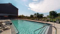 Located at 4125 Hayden Lofts Place in Dublin, Ohio, this single-level amenity space is part of the Hayden Lofts community and is designed to provide residents with a premier outdoor retreat. The centerpiece of the space is a large, resort-style swimming pool featuring crystal-clear water and a broad surrounding deck ideal for relaxation and recreation. The pool deck is well-appointed with multiple lounge chairs arranged to take full advantage of the open sky and scenic tree-lined views beyond the perimeter fencing. A sleek iron fence encloses the area, offering both security and an unobstructed sightline to the surrounding landscape. The adjacent building features a bold, modern exterior with dark metal cladding, contributing to the contemporary aesthetic of the overall community. The outdoor area is thoughtfully designed to accommodate both active use and leisurely lounging, creating an inviting atmosphere throughout the warmer months. The combination of clean architectural lines, generous open space, and natural surroundings gives this amenity area a polished yet relaxed character that is well-suited to the Hayden Lofts community in Dublin, OH.
