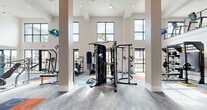 Located at 3280 Champion Ring Rd in Fort Myers, Florida, this two-story fitness center offers a generously sized, fully equipped workout facility spread across two floors. The main level commands attention with soaring double-height ceilings, large floor-to-ceiling windows, and glass doors that flood the space with natural light and provide direct views of the outdoor pool and surrounding grounds. Structural columns punctuate the open layout, lending an architectural rhythm to the expansive floor plan. The ground floor is outfitted with a wide selection of strength and functional training equipment, including cable machines, a multi-functional rig, plate-loaded machines, a punching bag, benches, and a robust dumbbell rack. A dedicated functional training zone features kettlebells, medicine balls, resistance tools, and a rowing machine, all set against clean white walls with colorful area rugs defining workout zones. A staircase connects to the upper level, where a mezzanine overlooks the main floor below. The second floor houses a row of treadmills and ellipticals positioned beside windows that frame outdoor views, along with additional cardio equipment. Consistent carpet tile flooring in blue, teal, and orange tones carries throughout both levels, reinforcing a modern, energetic aesthetic.