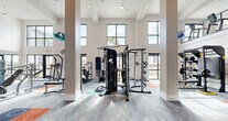 Located at 3280 Champion Ring Rd in Fort Myers, Florida, this two-story fitness center offers a generously sized, fully equipped workout facility spread across two floors. The main level commands attention with soaring double-height ceilings, large floor-to-ceiling windows, and glass doors that flood the space with natural light and provide direct views of the outdoor pool and surrounding grounds. Structural columns punctuate the open layout, lending an architectural rhythm to the expansive floor plan. The ground floor is outfitted with a wide selection of strength and functional training equipment, including cable machines, a multi-functional rig, plate-loaded machines, a punching bag, benches, and a robust dumbbell rack. A dedicated functional training zone features kettlebells, medicine balls, resistance tools, and a rowing machine, all set against clean white walls with colorful area rugs defining workout zones. A staircase connects to the upper level, where a mezzanine overlooks the main floor below. The second floor houses a row of treadmills and ellipticals positioned beside windows that frame outdoor views, along with additional cardio equipment. Consistent carpet tile flooring in blue, teal, and orange tones carries throughout both levels, reinforcing a modern, energetic aesthetic.