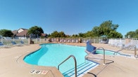 This single-story amenity space offers residents access to two distinct recreational areas designed for both relaxation and active living. The outdoor pool area is a standout feature, showcasing a generously sized swimming pool with crystal-clear water, a shallow entry point marked at three feet, and an adjacent spa finished in blue tile. The pool deck is surrounded by a white perimeter fence and lined with lounge chairs, providing a comfortable setting for outdoor leisure. Mature trees border the space, offering natural shade and a green backdrop under open skies.

The indoor fitness center is thoughtfully organized and outfitted with a comprehensive selection of equipment. A vibrant teal accent wall featuring athletic silhouette graphics energizes the space and reinforces its purpose. Cardio equipment includes multiple treadmills, elliptical trainers, and a stationary bike, while a Precor multi-station strength machine supports full-body resistance training. A wall-mounted television, wall clock, and built-in cabinetry add functional touches throughout. The fitness area is carpeted, well-lit with overhead fluorescent lighting, and accessible via a glass-panel entry door, creating a clean and practical environment suited for regular use.