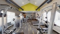 This fitness center offers a comprehensive workout environment designed to support a wide range of exercise routines. The space is defined by striking vaulted ceilings accented with a bold yellow interior peak, giving the room a sense of openness and energy. Exposed industrial-style ductwork adds a modern edge to the overall aesthetic, while recessed lighting ensures the space is well-lit throughout.

The floor plan is thoughtfully organized, with patterned carpet tile — featuring a graphic yellow design — covering the main workout area and transitioning to hardwood-style flooring in a dedicated stretching and free weights zone. Full-length mirrors line one wall, enhancing the sense of space and allowing users to monitor their form during exercise.

The cardio section is equipped with treadmills, elliptical machines, and stair climbers, all positioned near windows with horizontal blinds that invite natural light. The strength training area includes a selection of Precor cable and resistance machines, a Smith machine with a bench, and a full dumbbell rack. A wall-mounted television and a motivational "beastmode focus" mirror decal contribute to an engaging atmosphere. Multiple windows throughout the space provide views to the exterior, keeping the environment feeling fresh and connected to the outdoors.