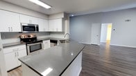 This single-floor space showcases a clean, modern aesthetic with an open-concept layout that seamlessly connects the kitchen and living areas. The kitchen is a standout feature, equipped with stainless steel appliances including a range, microwave, and dishwasher. Crisp white cabinetry with flat-panel doors lines the walls, complemented by a subtle tile backsplash that adds texture without overwhelming the space. A large kitchen island anchors the room, topped with a polished grey quartz countertop that provides ample workspace and features an undermount stainless steel sink. The flooring throughout is a warm-toned wood-look plank, offering both durability and visual continuity as it flows from the kitchen into the open living area. The walls are painted in a soft, cool grey tone that enhances the bright, airy feel of the space. Recessed and overhead lighting ensures the kitchen is well-lit, while the open living area beyond benefits from the overall brightness of the unit. A white paneled door leads to an additional room, suggesting a versatile floor plan. The overall ambiance is contemporary, clean, and move-in ready.