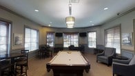 This single-floor recreational lounge is designed for social gathering and entertainment, combining a polished aesthetic with practical amenities. At the heart of the space sits a full-size billiards table with a rich wood finish, positioned beneath a statement tiered drum pendant light that anchors the room. Recessed lighting throughout ensures the space is well-lit at all times.

The room features an octagonal or multi-angled layout, with walls lined by a series of large windows dressed with horizontal blinds, allowing generous natural light to fill the interior while maintaining privacy. The neutral gray-toned walls and warm wood-look flooring create a welcoming and contemporary ambiance.

Multiple flat-screen televisions are mounted on the walls, making this space ideal for watching sports or other entertainment alongside games. Seating is thoughtfully distributed throughout, including high-top pub tables with bar-height chairs clustered near the windows, as well as a plush upholstered armchair in the corner for more relaxed lounging.

The overall design strikes a balance between casual comfort and refined style, offering a versatile environment suitable for socializing, gaming, and relaxation. The combination of natural light, layered artificial lighting, and well-chosen furnishings makes this lounge an inviting and functional communal space.
