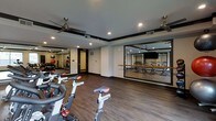 This single-floor fitness facility is thoughtfully designed to support a range of workout activities. The main space is anchored by a row of stationary cycling bikes arranged on rich, dark hardwood-style flooring, creating a dedicated cycling studio atmosphere. Ceiling fans with sleek, modern finishes are mounted overhead, ensuring air circulation and comfort during intense sessions.

Large mirrored wall panels define the space, visually expanding the room while allowing users to monitor their form. The mirrors reflect an adjacent weight and strength training area, which is visible through a wide mirrored opening and features a wall-mounted rack of medicine balls and additional equipment. A ballet-style barre runs along one mirrored wall, suggesting the space also accommodates stretching or barre-style classes.

Recessed lighting is integrated throughout the ceiling, providing bright, even illumination across the workout areas. Dark baseboards and trim offer a clean contrast against the neutral walls, lending the space a polished, modern aesthetic. A wall-mounted display screen in the cycling area adds a tech-forward touch, ideal for guided classes or entertainment during workouts.