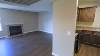 This single-story space offers a straightforward, open layout with a seamless flow between the living area and kitchen. The living room is generously sized, featuring dark hardwood-style flooring that runs throughout and provides a warm, grounding contrast to the neutral beige walls. White baseboards frame the perimeter cleanly, adding a crisp finish to the space. A fireplace with a simple mantle serves as a subtle focal point along one wall, offering both visual interest and functional appeal. The textured ceiling adds dimension overhead, while recessed ventilation is visible, suggesting a functional HVAC system. A half-wall partition separates the kitchen from the main living area, providing a degree of definition without fully closing off the space. The kitchen features dark cabinetry and a light-toned countertop, visible from the transition point between rooms. Light switches are conveniently positioned at the partition wall. The overall ambiance is clean, understated, and move-in ready, with neutral tones and durable finishes that provide a versatile backdrop suitable for a variety of interior styles.