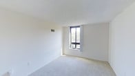 This single-floor space presents a clean and minimalist interior defined by neutral tones throughout. The room is finished with wall-to-wall light beige carpeting that contributes to a quiet, comfortable atmosphere underfoot. Crisp white walls and white baseboards create a bright, open feel, while a textured white ceiling adds subtle visual interest overhead. A centrally positioned, floor-to-height window serves as the focal point of the room, framed in dark contrasting trim and fitted with a sheer white curtain. The window allows generous natural light to fill the space while offering a pleasant view of mature trees outside, suggesting a verdant surrounding environment. A wall-mounted HVAC vent is visible near the ceiling, indicating a built-in climate control system. The overall layout is open and unobstructed, providing flexibility for various furniture arrangements. The space maintains a simple, functional aesthetic with no ornamental detailing, making it highly adaptable to different interior styles. The combination of natural light, neutral finishes, and soft carpeting gives the room a calm and airy ambiance.