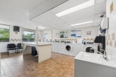 Located at 350 Elliott St in Windsor, Ontario, this shared laundry facility is a well-organized, single-floor space designed to serve the needs of its users efficiently. The room features a row of top-loading washers along one wall, complemented by a bank of stacked dryer units, providing ample capacity for multiple users at once. Front-loading washers are also available, offering a variety of machine options to suit different laundry needs. The machines are equipped with modern card-based payment terminals, eliminating the need for coins and streamlining the laundry experience. Informational signage regarding proper washer and dryer usage is posted throughout the room, keeping users well-informed. The space is finished with durable tile flooring in a warm earth tone, white walls accented by a neutral tile border, and a drop ceiling with recessed and overhead lighting that keeps the room bright and functional. A large folding table provides a practical surface for sorting and folding laundry. Seating is available for users waiting on their loads. Windows along one wall allow natural light to enter, adding to the overall brightness of the space. A portable fan and additional cleaning equipment are on hand to maintain comfort and cleanliness. The layout is open and efficient, making the most of the available floor area.