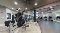 Located at 2800 Pontiac Dr in Aurora, IL, this modern fitness center offers a comprehensive workout environment across a single, thoughtfully designed floor. The space is divided into distinct functional zones, each catering to different aspects of physical training.

The strength training area is anchored by a multi-station cable machine and a variety of resistance equipment, all set on patterned carpet flooring. Large wall mirrors amplify the sense of space and allow for form monitoring during workouts. Dark structural columns punctuate the layout, adding an industrial-contemporary aesthetic that is complemented by a combination of recessed lighting and stylish pendant fixtures overhead.

The cardio section is generously outfitted with treadmills, ellipticals, upright bikes, and stair climbers — all featuring modern digital consoles. Tall windows along the perimeter flood this zone with natural light and offer views of the surrounding neighborhood, creating an open and energizing atmosphere. Ceiling fans throughout ensure comfortable airflow during intense sessions.

A dedicated open-floor functional training area includes a mounted interactive display screen, a freestanding punching bag, and a storage unit stocked with accessories such as resistance bands and stability equipment. A water fountain station, accented by a bold navy feature wall with curated artwork, provides a convenient hydration stop. An adjacent locker room rounds out the facility's amenities.