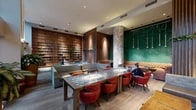 This thoughtfully designed two-floor amenity space blends a refined social atmosphere with practical functionality. The main floor is anchored by a dramatic double-height atrium flooded with natural light through expansive floor-to-ceiling windows, offering views of a lush outdoor terrace landscaped with bamboo and potted greenery. A striking floor-to-ceiling bookshelf wall fitted with wooden drawers and curated book collections serves as a defining architectural centerpiece, complemented by warm wood-paneled columns and a bold emerald chalkboard accent wall. Seating throughout is eclectic yet cohesive, featuring tufted sectional sofas, red leather armchairs, patterned communal tables, and intimate leather club chairs arranged around round side tables. A second lounge area on the same level offers a quieter retreat with patterned tile walls, ambient cove lighting, and a glass coffee table set atop a textured area rug. The second floor hosts an open co-working and recreation area with long communal tables, task seating, a pool table, and additional workspaces positioned near windows with outdoor views. A compact kitchenette services the space, equipped with a Nespresso machine, beverage station, sink, and induction cooktop, all finished with wood cabinetry and patterned tile flooring. Private nook spaces with individual climate control provide focused work areas throughout.