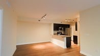 This single-floor residence showcases a thoughtfully designed open-concept layout that seamlessly connects the living and kitchen areas. The space is grounded by warm hardwood flooring that runs throughout, adding a sense of continuity and warmth to the interior.

The kitchen is a standout feature, equipped with stainless steel appliances including a refrigerator, microwave, and range. Rich dark cabinetry provides a striking contrast against lighter countertops and walls, while a peninsula with a granite-style countertop offers additional prep space and casual seating. Track lighting is installed in both the kitchen and living areas, providing versatile and well-distributed illumination.

The open living area benefits from the flowing layout, offering generous usable space and a clean, unfurnished canvas that allows for flexible furniture arrangements. Neutral, warm-toned walls and white trim complement the overall aesthetic, maintaining a bright and inviting ambiance. A secondary kitchen area with lighter cabinetry is also visible, hinting at additional storage and workspace. The combination of modern finishes, practical design, and cohesive materials makes this a functional and visually appealing single-floor residence.