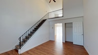 This two-story space makes a strong first impression with its dramatically vaulted ceilings that rise to a striking height in the main living area, creating an expansive, airy atmosphere. A carpeted staircase with sleek black metal railing ascends to the upper level, where an open landing overlooks the main floor below, adding to the sense of openness and vertical scale. A ceiling fan is mounted at the peak, providing both function and visual interest at height.

The main level features warm hardwood-style flooring that extends throughout the open living space, contributing a clean and cohesive aesthetic. The walls are finished in crisp white, enhancing the brightness of the interior. Natural light filters in through vertical blinds visible in an adjoining room, adding warmth to the overall ambiance.

A doorway from the main living area leads to an additional room, which includes a built-in closet with shelving, offering practical storage. A white panel door nearby provides further access to additional spaces. The layout balances openness with defined functional areas, making it a versatile and well-structured two-level space.