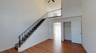 This two-story space makes a strong first impression with its dramatically vaulted ceilings that rise to a striking height in the main living area, creating an expansive, airy atmosphere. A carpeted staircase with sleek black metal railing ascends to the upper level, where an open landing overlooks the main floor below, adding to the sense of openness and vertical scale. A ceiling fan is mounted at the peak, providing both function and visual interest at height.

The main level features warm hardwood-style flooring that extends throughout the open living space, contributing a clean and cohesive aesthetic. The walls are finished in crisp white, enhancing the brightness of the interior. Natural light filters in through vertical blinds visible in an adjoining room, adding warmth to the overall ambiance.

A doorway from the main living area leads to an additional room, which includes a built-in closet with shelving, offering practical storage. A white panel door nearby provides further access to additional spaces. The layout balances openness with defined functional areas, making it a versatile and well-structured two-level space.