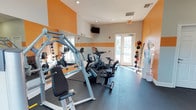 This fitness center is designed to provide a functional and motivating workout environment. The space is characterized by bold orange accent walls paired with crisp white ceilings and trim, creating an energetic atmosphere conducive to exercise. Durable rubber flooring covers the workout area, offering both safety and comfort underfoot.

The room is outfitted with a variety of exercise equipment to support diverse fitness routines. Cardio options include multiple stationary and spin-style cycling machines, while a multi-function strength training machine provides options for resistance and weight-based exercises. A medicine ball rack along one wall offers additional functional fitness tools, and a water cooler ensures convenient hydration.

A wall-length mirror runs along one side of the room, visually expanding the space while allowing users to monitor their form during workouts. Natural light filters in through windows and a set of glass-paned double doors, which also provide direct outdoor access. A wall-mounted flat-screen television is positioned for visibility from multiple workout stations, adding an entertainment option during sessions. Recessed lighting supplements the natural light, keeping the space bright and well-illuminated. Overall, the fitness center delivers a practical, well-appointed environment suited for a well-rounded workout.