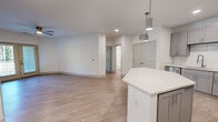 This well-appointed single-floor residence in Carmel, Indiana offers a thoughtfully designed open-concept layout with two bedrooms, two bathrooms, and two closets. The combined kitchen and living area forms the heart of the home, showcasing light-toned hardwood-style flooring and a clean, neutral color palette throughout. The kitchen is a standout feature, equipped with grey shaker-style cabinetry, white quartz countertops, a subway tile backsplash, stainless steel appliances including a range, over-the-range microwave, dishwasher, and refrigerator, plus a hexagonal center island with pendant lighting above. The open living area flows seamlessly from the kitchen and features a distinctive decorative wall with a paneled grid accent, adding architectural character to the space. Ceiling fans are present in multiple rooms for added comfort. Both bedrooms offer plush carpeting, natural light through double windows with views to the outdoors, and ceiling fans. Each bathroom is finished with wood-look tile flooring, white subway tile tub surrounds with brushed nickel fixtures, and clean white vanities. Two dedicated closets provide practical storage. The residence presents a cohesive, modern aesthetic ideal for comfortable everyday living in the heart of Carmel, IN.