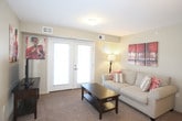 This single-floor residence offers a thoughtfully organized layout designed for comfortable everyday living. The living room serves as the central gathering space, finished with neutral-toned carpet and warm wall colors. It features a sofa, accent chairs, and decorative artwork, and benefits from natural light streaming through glass patio doors that open to the outdoors—an inviting architectural detail that enhances the room's openness.

The kitchen is efficiently designed with abundant warm oak cabinetry providing ample storage, laminate countertops, a double-basin sink, and white appliances including an electric range and dishwasher. Light tile flooring completes the practical, easy-to-maintain space.

The primary bedroom is a comfortable retreat, featuring plush carpeting, a double closet with bifold doors, and soft ambient lighting. A second bedroom similarly offers carpeted flooring, a window with natural light, and space for a bed and desk. An additional flexible room, also carpeted and windowed, functions well as a guest bedroom or home office.

The full bathroom includes a warm-toned vanity with wood cabinetry, a white countertop, a wall-mounted mirror, a toilet, and a shower-tub combination enclosed by a striped fabric curtain, all set on light tile flooring.