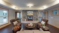 Located at 568 Settlers Ridge Pkwy in Woodbury, Minnesota, this single-floor communal space blends comfort and modern elegance into a cohesive, inviting environment. The lounge area serves as the heart of the space, anchored by a striking floor-to-ceiling stacked stone fireplace that creates a warm, welcoming focal point. Plush leather wingback chairs and upholstered seating are arranged thoughtfully around a round wood coffee table, encouraging conversation and relaxation. Recessed lighting and a statement flush-mount ceiling fixture illuminate the room, while large windows allow natural light to filter in, brightening the neutral-toned walls accented with warm wood trim. Dark hardwood flooring adds a layer of sophistication throughout. The kitchen and entertaining area is equally impressive, featuring an expansive granite-topped island with dark wood cabinetry and sleek hardware. Glass pendant lights hang overhead, casting a warm glow over the prep and gathering space. Stainless steel appliances, including a full-size refrigerator, complement the dark herringbone accent wall behind the kitchen station, where a wall-mounted television adds a social entertainment element. French doors at the rear of the kitchen open to an outdoor seating area, connecting the interior seamlessly to the outside. The overall ambiance is refined yet approachable, designed for both casual gatherings and community events.