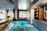 Located at 925 W 20th St in Houston's vibrant Heights neighborhood, this thoughtfully designed single-floor space blends contemporary aesthetics with functional amenity-style living. The open-plan layout is anchored by a striking teal patchwork carpet that flows through the main lounge area, creating a vivid visual centerpiece beneath recessed lighting and a sleek dark ceiling.

The lounge features a built-in banquette seating nook framed by a recessed alcove, accented with bold pink throw pillows and decorative circular wall art, offering an intimate gathering spot. Slender pedestal side tables complement the seating arrangement, while a large window allows natural light to filter in, providing views of the surrounding streetscape.

A dedicated wet bar area showcases warm wood cabinetry with gold hardware, a granite countertop, and a full mirrored tile backsplash that amplifies light and depth throughout the space. Open wooden shelving displays curated décor, and an under-counter refrigerator adds practical utility. A herringbone-patterned hardwood floor defines this bar zone, contrasting elegantly with the teal carpet of the lounge.

A built-in shelving unit with display cases and a low stone-topped credenza lines one wall, offering both storage and display for artwork and accessories. The overall ambiance is sophisticated, energetic, and well-appointed, ideal for social gatherings and community engagement.