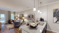This single-floor residence offers a thoughtfully designed open-concept layout that seamlessly connects the kitchen, dining, and living areas. The kitchen is outfitted with dark espresso cabinetry, granite countertops, a subway tile backsplash, and a full suite of stainless steel appliances, including a refrigerator, range, microwave, and dishwasher. A large kitchen island with an undermount sink and pendant lighting serves as the central hub between the kitchen and the living space. Crown molding and rich hardwood-style flooring flow throughout the main living areas, lending a polished, cohesive aesthetic. The living room is generously sized, featuring large windows that invite natural light and provide access to an exterior door. A dedicated dining area is defined by an elegant chandelier and ample space for entertaining. The two bedrooms are carpeted for comfort, each featuring ceiling fans and windows with views to the outside. The primary bathroom offers dark cabinetry with granite countertops and a tiled shower-tub combination, while the secondary bathroom mirrors a similarly clean and functional design with tile flooring. Neutral tones in warm greige carry throughout, creating a calm and inviting atmosphere across every room.