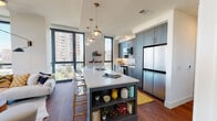 This single-floor residence showcases a thoughtfully designed open-concept layout with contemporary finishes and generous natural light throughout. Floor-to-ceiling windows frame urban skyline views, creating a bright and airy atmosphere across the living spaces.

The kitchen serves as a central focal point, featuring slate-blue cabinetry with brushed gold hardware, a white quartz waterfall island with built-in shelving, and stainless steel appliances. Globe pendant lights with brass accents hang overhead, while a marble-style backsplash adds visual texture. Acrylic and gold barstools line the island, making it ideal for casual dining.

The bedrooms are calm and well-appointed. The primary suite includes expansive windows with sheer drapery, a ceiling fan, soft carpeting, and access to a walk-in closet with a window. A secondary bedroom features large floor-to-ceiling windows, plush carpet, and a seating area.

The bathroom presents a navy blue vanity with gold drawer pulls, white quartz countertop, and light tile flooring. A hallway connects the rooms with smooth transitions between wood and tile surfaces. Throughout the residence, a cohesive palette of neutral tones, warm wood accents, and modern fixtures establishes a polished, urban aesthetic.