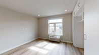 This single-floor unit offers a clean, modern design characterized by neutral warm-beige walls, light wood-grain flooring, and recessed lighting throughout. The main living area is open and airy, benefiting from a large window that invites abundant natural light and provides views of the surrounding urban cityscape. A wall-mounted PTAC heating and cooling unit ensures year-round climate comfort. Built-in cabinetry with open shelving provides practical storage and display space within the living area.

The kitchen is compact yet fully functional, featuring white flat-panel cabinetry that extends to the ceiling for ample storage. Stainless steel appliances include a full-size refrigerator, dishwasher, electric range with oven, and an over-the-range microwave, all complemented by a stainless steel sink and sleek countertops.

The bathroom is finished with white subway tile in a generously sized, barrier-free walk-in shower equipped with a rainfall showerhead and handheld fixture, as well as brushed nickel grab bars — reflecting a thoughtful, accessibility-conscious design. The floor is finished in neutral tile, contrasting cleanly with the lighter flooring found throughout the rest of the unit. Overall, the space presents a bright, move-in-ready environment with a cohesive modern aesthetic.