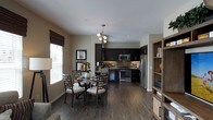 This single-floor space showcases an open-concept layout that seamlessly integrates the dining area and kitchen into one cohesive living environment. The kitchen is outfitted with dark espresso cabinetry that extends to the upper walls, complemented by a striking mosaic tile backsplash and a full suite of stainless steel appliances, including a French door refrigerator, a gas range, a built-in microwave, and a wine cooler. Under-cabinet lighting adds a warm, ambient glow to the workspace. Rich, dark hardwood-style flooring runs throughout, anchoring the space with a sense of warmth and continuity. The dining area features a round table surrounded by lattice-back chairs beneath a multi-bulb pendant light fixture, creating an inviting gathering spot. Natural light filters in through a large window dressed with dark Roman shades. Neutral warm-toned walls provide a clean backdrop that highlights the darker finishes. An open shelving unit on one side adds functional storage and display space, rounding out the practical yet stylish character of this well-appointed interior.