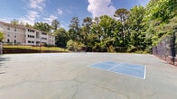 Located at 700 Summit Place Dr in Sandy Springs, Georgia, this outdoor recreational facility features a well-defined basketball court and an adjacent tennis court, both surfaced in concrete and enclosed by sturdy black chain-link fencing. The basketball court is marked with painted court lines and key areas, while the tennis court displays clearly visible boundary lines. Courtside seating in the form of wooden bleachers provides a spot for spectators to observe play. Overhead lighting poles are positioned throughout the facility, extending usability into evening hours. The courts are set against a backdrop of mature, densely wooded surroundings, with tall deciduous and evergreen trees providing a natural canopy and a sense of seclusion. Tiered brick retaining walls and manicured grassy slopes frame one side of the facility, leading up to a multi-story residential building with a contemporary design featuring large windows and balconies. A white split-rail fence lines the upper perimeter, adding a classic touch to the landscape. The overall setting combines active recreational amenities with a lush, park-like environment, making it a practical and visually appealing outdoor space for residents and visitors alike.