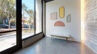This single-floor commercial space exemplifies contemporary architectural design, blending raw industrial materials with refined finishes to create a polished and welcoming environment. Polished terrazzo-style concrete floors run throughout, reflecting light and lending the space a clean, sophisticated look. The entryway makes an immediate impression with dramatic floor-to-ceiling windows framed by exposed board-formed concrete columns, flooding the interior with abundant natural light and offering street-level views of the surrounding urban landscape. A sleek white upholstered bench with brass legs sits beneath a curated wall installation featuring geometric framed panels in soft pastel tones — yellow, blush, and sage — adding an artistic focal point to the reception area. The corridors are wide and well-lit, with crisp white walls, handrails, and recessed ceiling lighting that guide movement through the space. A dark-painted ceiling accent in select areas adds depth and architectural contrast. Keycard-access black doors and a stainless steel elevator underscore the building's modern infrastructure. The layout flows logically from the light-filled entry through functional corridors toward additional rooms, making this space well-suited for professional, creative, or wellness-oriented commercial use.