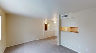 This single-story space offers a straightforward, functional layout designed for comfortable everyday living. The main living area features wall-to-wall carpet in a neutral tone, providing a soft underfoot feel that complements the warm, beige wall palette throughout. A pass-through kitchen counter with a tile backsplash connects the kitchen to the living space, allowing for an open, communicative flow between the two areas. White cabinetry is visible above the pass-through, contributing to a clean and bright aesthetic. An HVAC vent is positioned overhead near the kitchen transition, ensuring efficient climate control. Adjacent to the main living area, a secondary room is visible through a wide opening framed by a decorative ledge, distinguishing the two spaces while maintaining visual connectivity. This adjoining room features wood-look hard flooring and is fitted with a ceiling fan, adding both practicality and airflow. The consistent neutral color scheme, smooth white ceilings, and open sightlines create an airy, cohesive atmosphere throughout the space.