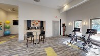 Located at 3190 Skinner Mill Rd in Augusta, Georgia, this single-floor fitness center is designed to offer a comprehensive and welcoming workout environment. The space features soaring ceilings that create an open, airy atmosphere, complemented by neutral gray and white walls accented with bold navy blue trim that adds a modern, polished aesthetic throughout.

The fitness floor is finished with durable, multi-toned carpet tiles in gray and yellow, providing both visual interest and a practical surface for exercise. Large windows and glass doors flood the space with natural light while offering views of the surrounding outdoor area, enhancing the overall sense of openness.

The facility is well-equipped with a wide range of exercise options. Cardio equipment includes treadmills, elliptical machines, and a stationary cycling bike. A cable machine with a weight bench and a full dumbbell rack support strength training, while resistance bands, medicine balls, exercise mats, and balance equipment accommodate functional and flexibility training.

A dedicated refreshment nook, finished in navy cabinetry and accented with colorful artwork, features a coffee station and bar-height seating — offering a convenient spot to recharge before or after a workout. Glass interior doors connect the fitness center to an adjacent lounge or common area, reinforcing the facility's integration within a larger community setting.