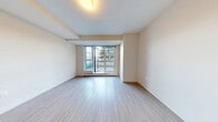 This efficiently designed single-floor unit offers a well-organized layout accommodating two kitchen areas, a living room, one bathroom, and a dedicated laundry space within a compact footprint. The interior presents a clean, contemporary aesthetic, characterized by light wood-effect flooring, neutral cream-toned walls, and recessed ceiling details that add subtle architectural interest throughout.

The living room is notably bright, benefiting from a large floor-to-ceiling sliding glass door and adjacent window that frame views of surrounding greenery and provide direct access to a private outdoor patio. The patio, accessible via a glass-paneled entrance door, offers an outdoor extension of the living space with stone tile flooring.

The kitchen features a streamlined, modern design with flat-front grey cabinetry, white countertops, a stainless steel sink with a contemporary pull-down faucet, an induction cooktop, under-cabinet lighting, and overhead stainless steel range hood, complemented by upper cabinetry extending to the ceiling for maximum storage.

The bathroom is finished with large-format tile flooring, a floating grey vanity with a white basin, a full-width mirror, and a soaking bathtub with tiled surround. A separate laundry room with wood-effect flooring and built-in hookups completes the functional layout of this neatly appointed space.
