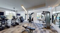 Located at 4027 Watercraft Ferry Avenue in Wilmington, NC, this fitness center is a thoughtfully designed, single-floor facility built to support a comprehensive workout experience. The main exercise area is expansive and airy, featuring soaring ceilings, large multi-pane windows that flood the space with natural light, and views of the surrounding community. The flooring throughout the main gym area consists of a stylish checkered tile pattern that adds a modern aesthetic while providing durability underfoot. Equipment includes elliptical machines, treadmills, cable machines, TRX suspension trainers, a cable crossover system, adjustable benches, and a full dumbbell rack, catering to a wide range of fitness needs. Ceiling fans and mounted flat-screen televisions add comfort and entertainment throughout the space. A separate spin studio offers a clean, light-filled room with hardwood-style flooring, multiple cycling bikes, and a wall-mounted stability ball rack, creating an ideal environment for group or individual cycling sessions. A dedicated entryway area features built-in cabinetry, an informational bulletin board, and a wall-mounted water fountain for convenience. Accent wallpaper detailing and recessed lighting complete the polished, contemporary ambiance of this well-equipped fitness facility.