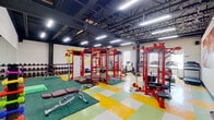 This dynamic fitness facility offers a comprehensive workout environment designed to energize and inspire. The open, single-floor layout provides generous training space, with soaring ceilings that expose industrial-style ductwork and structural elements, all illuminated by rows of bright overhead linear lighting that ensure excellent visibility throughout.

The strength training area is a standout feature, anchored by bold red multi-functional power rigs and cable machines. A full dumbbell rack lines one wall, offering a wide range of free weights alongside benches, medicine balls, battle ropes, and resistance accessories. Dedicated lifting platforms with rubber matting provide safe and functional workout zones.

The cardio section is positioned near a bank of large windows fitted with zebra-style blinds, allowing natural light to flood the space. Multiple treadmills and a stationary bike are neatly arranged in this area, creating a bright and airy feel conducive to endurance training.

Large, vibrant pop-art murals decorate the walls, adding a bold, energetic aesthetic that complements the facility's active atmosphere. The colorful checkerboard flooring in green, yellow, and white further enhances the lively character of the space. Full-length mirrors amplify the sense of openness, making the facility feel expansive. Overall, this gym delivers a motivating, well-rounded environment suitable for a wide variety of fitness goals.