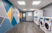 Located at 245 Atlantic Ave in Long Branch, NJ, this single-floor laundry facility combines practical functionality with a clean, modern aesthetic. The space is designed for efficiency and ease of use, accommodating multiple stacked dryer units and a row of front-load washing machines, all equipped with digital payment interfaces for convenient, cashless operation.

The interior is finished with light wood-look tile flooring throughout, lending warmth to the otherwise utilitarian space. The drop ceiling is fitted with recessed panel lighting that provides consistent, bright illumination across the entire room. Walls are painted in a cool gray tone, offering a neutral and tidy backdrop.

A standout design element is the bold geometric mural adorning one wall, featuring angular shapes in teal, yellow, gray, and white — adding a vibrant, contemporary character to the room. Natural light filters in through windows positioned near the entry door, offering views of the surrounding greenery outside.

The layout allows for ample open floor space between the machines and the opposite wall, making it easy for multiple users to move freely. A payment kiosk station is also present within the space. The facility is accessible via a standard entry door with an automatic door closer, ensuring a straightforward and welcoming experience for all users.